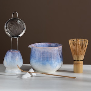 Könnte beinhalten: Ein Matcha-Teeset bestehend aus einer Keramikschale mit blau-weißem Farbverlauf, einem Bambusbesen, einem Bambuslöffel auf einer weißen Keramikablage und einem Metallsieb auf einem passenden blau-weißen Keramikständer.