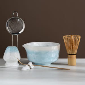 Puede incluir: Un juego de té matcha que incluye un cuenco y un soporte para batidor de cerámica esmaltada cristalina en blanco y azul claro, un batidor de bambú, una cuchara de bambú sobre un soporte blanco y un colador de acero inoxidable con soporte.