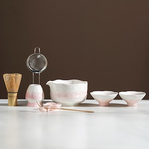 Op de afbeelding: Een matcha theeset met een bamboe klopper, een keramische kom, twee kleine kommen, een zeef, een keramische houder, een lepel en een schep. De set heeft witte en roze accenten, wat een serene esthetiek creëert.