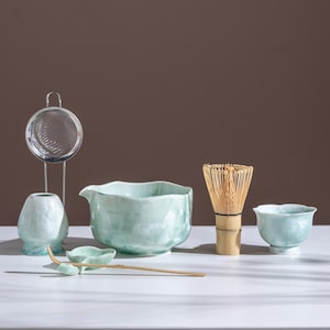 May include: A matcha tea set featuring a light blue ceramic bowl, whisk, small cup, scoop, strainer, and vase. The set is arranged on a white surface with a brown backdrop. The whisk is made of bamboo.