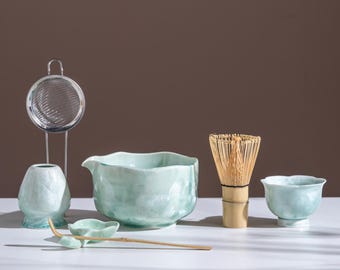 Op de afbeelding: Een matcha-theeset met een lichtblauwe keramische kom, een garde, een kleine kop, een schep, een zeef en een kleine vaas. De set is geplaatst op een wit oppervlak met een bruine achtergrond. De garde is gemaakt van bamboe.