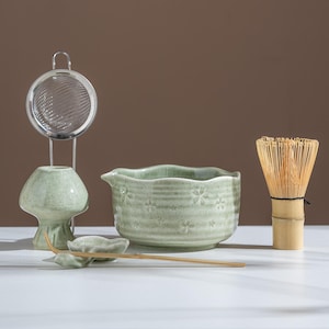 Könnte beinhalten: Ein Matcha-Teeset mit einer hellgrünen Keramikschale mit floralen Mustern, einem Bambusbesen, einem kleinen Keramik-Löffel, einem Keramikständer mit einem Edelstahl-Sieb und einem Bambuslöffel. Das Set steht auf einer weißen Oberfläche.
