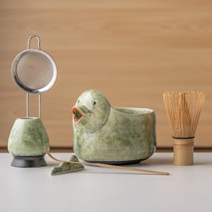 Może przedstawiać: Zestaw do herbaty matcha zawierający jasnozieloną ceramiczną miseczkę w kształcie kaczki z otwartym dziobem, pasujący zielony stojak na miotełkę z metalowym sitkiem, bambusową miotełkę oraz cienką bambusową łyżeczkę na małej ceramicznej podstawce.