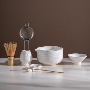 Op de afbeelding: Een matcha theeset bestaande uit een bamboe garde, een witte keramische houder, een metalen zeef, een witte keramische schenkkom met een textuur, een kleine witte kom en een lange, dunne bamboe theeschep op een houder.