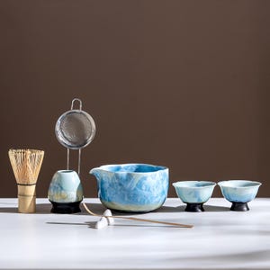 May include: A matcha tea set comprising a bamboo whisk, a ceramic bowl with a spout, two small tea bowls, a ceramic whisk holder, a metal sieve, and a bamboo scoop. The ceramic items have a blue and white marbled glaze, and the whisk is made of natural bamboo.