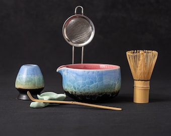 Può includere: Un set da tè matcha con una ciotola in ceramica blu e rosa, un frullino di bambù, un cucchiaino da tè, una tazza da tè, un portafrullino da tè e un colino da tè in acciaio inossidabile. La ciotola ha una superficie strutturata.