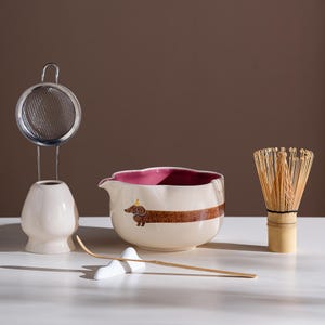 May include: A ceramic matcha tea set including a cream-colored bowl with a brown dachshund illustration wearing a yellow party hat, a pink interior, a bamboo whisk, a bamboo scoop on a white rest, and a metal sifter on a cream ceramic stand.