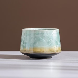 以下が含まれることがあります： 青と黄色の独特な結晶釉薬が施された陶器のカップ。カップは黒い台座と幅広で丸みを帯びた形状をしています。釉薬はテクスチャード加工されたひび割れ効果を生み出し、視覚的な面白さを加えています。カップはおそらく手作りです。