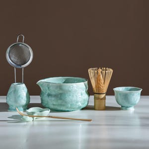 May include: A matcha tea set featuring a light green glazed ceramic bowl with a pouring spout, a matching whisk holder, a small tea bowl, a bamboo whisk, a bamboo tea scoop, a small ceramic rest, and a stainless steel mesh tea strainer.