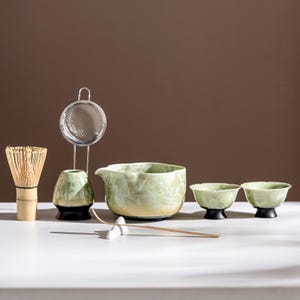 May include: A matcha tea set featuring a bamboo whisk, a metal sieve, a ceramic matcha bowl, two small tea bowls, and a bamboo scoop. The set has a green and cream color scheme with black accents. The items are arranged on a white surface.