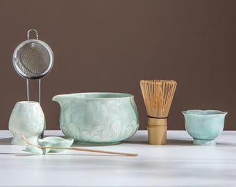 Puede incluir: Un juego de té matcha con un cuenco de cerámica azul claro, un batidor de bambú, una taza pequeña, un colador, una cuchara y un platillo. El juego está dispuesto sobre una superficie blanca contra un fondo marrón.