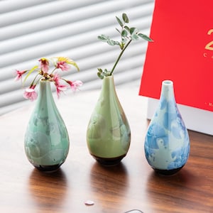 May include: Three ceramic bud vases in shades of green and blue, each with a unique floral pattern. The vases are filled with small sprigs of greenery and pink flowers. The vases are teardrop-shaped with a dark brown base.