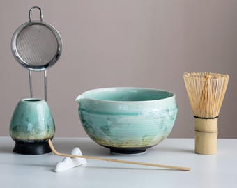 May include: A matcha tea set comprising a turquoise ceramic bowl, a bamboo whisk, a small ceramic vase, a stainless steel strainer, and a bamboo scoop. The bowl has a wide rim and a textured surface. The whisk is made of thin bamboo tines.