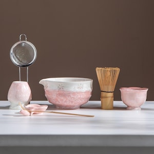 May include: A matcha tea set featuring a pink and white ceramic bowl, a bamboo whisk, a small pink bowl, a pink ceramic spoon, a pink ceramic holder, and a stainless steel strainer. The items are arranged on a white surface.