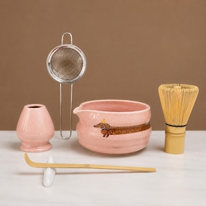 May include: A matcha tea set including a pink speckled ceramic bowl with a dachshund illustration, a matching pink whisk holder, a silver metal mesh strainer, a light-coloured bamboo whisk, and a long bamboo tea scoop resting on a white ceramic stand.