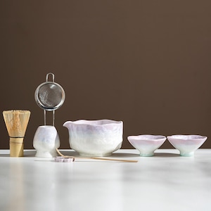 May include: A complete matcha tea set featuring a bamboo whisk, a stainless steel strainer, a ceramic bowl, two small tea bowls, and a bamboo scoop. The ceramic pieces have a pearlescent finish with pink and white hues.