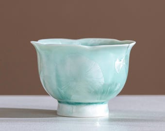 May include: A celadon-colored ceramic teacup with a flared, petal-like rim. The cup has a white base and a subtle, textured pattern on the exterior. The overall design is elegant and minimalist, suitable for serving tea or other beverages.