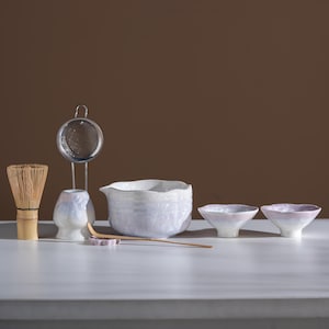 May include: A matcha tea set featuring a bamboo whisk, a metal tea strainer, a ceramic whisk holder, a large white and iridescent ceramic mixing bowl with a spout, a long bamboo scoop, and two small matching ceramic tea bowls with pink rims.
