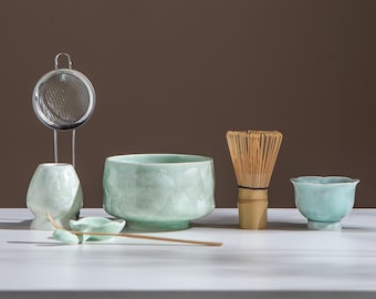 May include: A matcha tea set featuring a light green ceramic bowl, a bamboo whisk, a small tea cup, a ceramic spoon, a tea strainer, and a ceramic holder. The set is on a white surface with a brown backdrop.