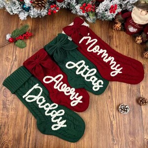 May include: Four knitted Christmas stockings in alternating red and dark green colors, each featuring a large bow at the top and white embroidered names. The stockings are labeled Mommy, Atlas, Avery, and Daddy in a cursive script font.