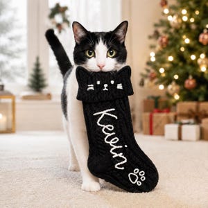 Op de afbeelding: Een zwart-witte kat die een zwarte kerstkous van kabelbreisel in de bek houdt. De kous heeft een kattengezichtje aan de bovenkant, de naam Kevin geborduurd in wit schrift en een kleine witte pootafdruk bij de onderste rand.