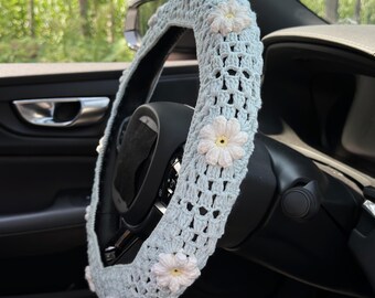 May include: A light blue crochet steering wheel cover adorned with white and yellow flower appliqués. The cover is designed to fit over a car's steering wheel, adding a decorative touch. The crochet pattern is open, creating a textured appearance.