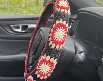 May include: A black, red, and cream crochet steering wheel cover. The cover features a repeating floral pattern. The steering wheel is inside a car with a black interior. The cover is designed to fit a standard size steering wheel.