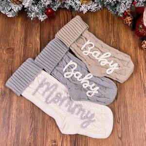May include: Four knitted Christmas stockings in beige, gray, and white, with the words "Baby" and "Merry" embroidered in white and gray. The stockings are on a wooden surface, with Christmas decorations in the background.