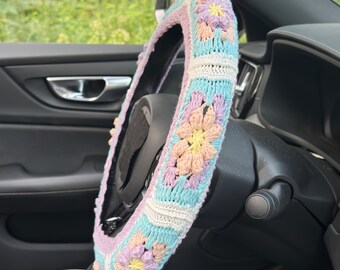 May include: A colorful, crocheted steering wheel cover. The cover features a floral pattern in pastel shades of pink, blue, yellow, and white. The cover is designed to fit a standard car steering wheel and adds a decorative touch to the vehicle's interior.