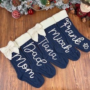 Puede incluir: Cinco calcetines navideños azul marino con lazos y puños blancos. Cada calcetín está personalizado con un nombre en escritura blanca: Mom, Dad, Tiana, Micah y Frank. El calcetín de Frank tiene una huella de pata.