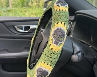 May include: Crocheted steering wheel cover in green, yellow, and gray. The cover features a repeating pattern of skulls and sunflowers. The cover is shown installed on a black steering wheel in a car.