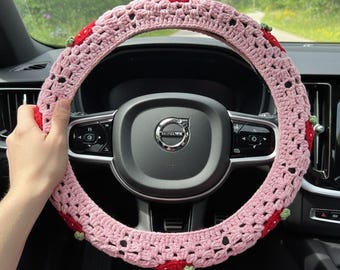May include: A pink crocheted steering wheel cover with a strawberry design. The cover is light pink with red strawberries and green leaves. The cover is on a black steering wheel in a car.