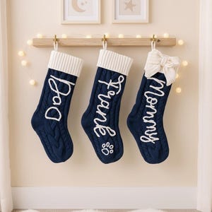 May include: Three navy blue cable knit Christmas stockings hang from a wooden shelf. Each stocking has a white cuff and white embroidered text: "Dog", "Frank", and "Mommy". The Frank stocking has a paw print.