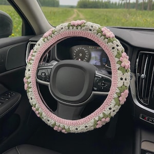 May include: A crocheted steering wheel cover in a car. The cover features a floral design with pink, white, and green accents. The steering wheel is black, and the car's dashboard is visible in the background.