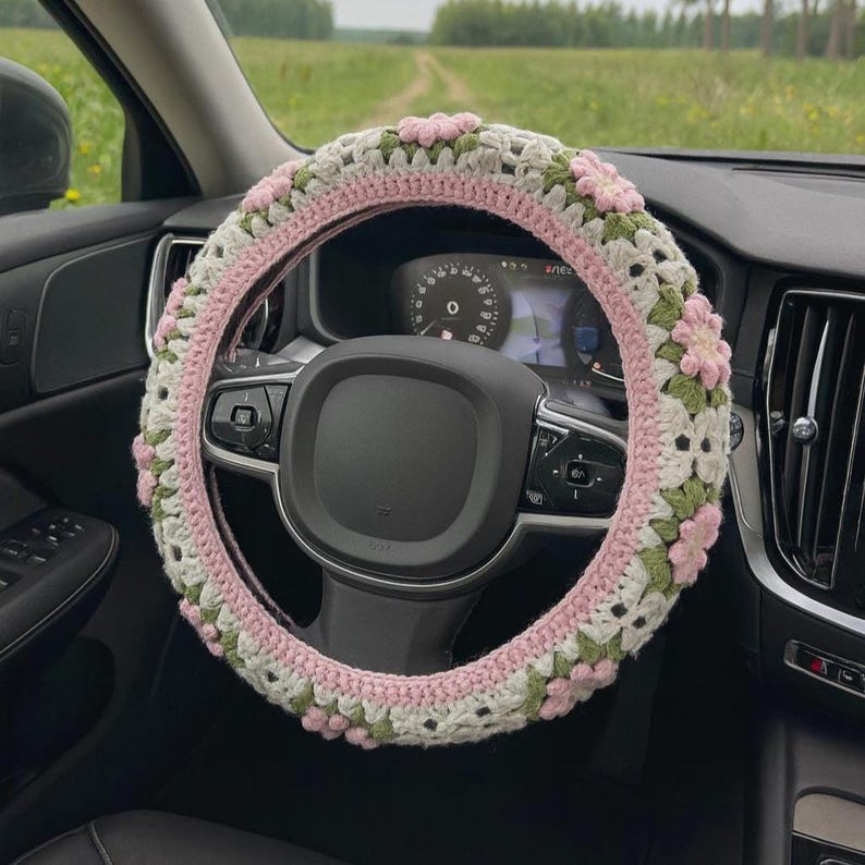 May include: A crocheted steering wheel cover in a car. The cover features a floral design with pink, white, and green accents. The steering wheel is black, and the car's dashboard is visible in the background.