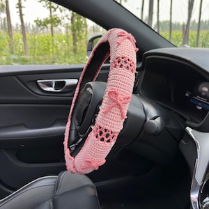 May include: A pink crocheted steering wheel cover with a decorative bow design. The cover is made of a soft, textured yarn and features openwork detailing. The cover is shown installed on a car's steering wheel.