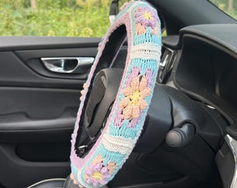 May include: A colorful, crocheted steering wheel cover. The cover features a floral pattern in pastel shades of pink, blue, yellow, and white. The cover is designed to fit a standard car steering wheel and adds a decorative touch to the vehicle's interior.