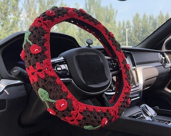 May include: A crocheted steering wheel cover in a car. The cover features a brown base with red accents and cherry designs. Green leaves and red cherries are crocheted onto the cover. The steering wheel is black.