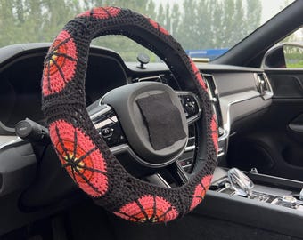 May include: A black crochet steering wheel cover with a repeating pattern of red and orange flower-like designs. The cover is installed on a black steering wheel inside a car.