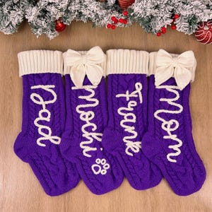 Pode incluir: Quatro meias de Natal em tricô trançado roxo com punhos canelados brancos. Cada meia apresenta um laço de crochê branco e letras cursivas brancas lendo Dad, Mochi com uma pata, Franke e Mom. As meias estão dispostas lado a lado.