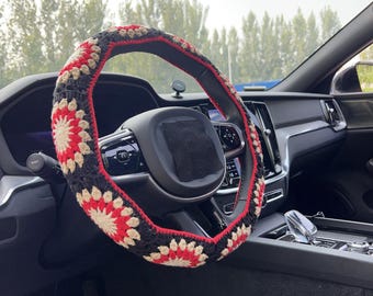 May include: A crocheted steering wheel cover in a car. The cover features a repeating floral pattern in red, black, and beige. The steering wheel is black, and the car's interior is also black.