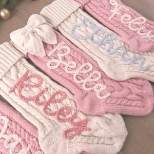Pode incluir: Uma coleção de meias de Natal em tricô com padrões de tranças em creme e rosa suave. Cada meia é personalizada com nomes bordados, incluindo Riley, Bella, Ethan e Felix, em cores de fio contrastantes.