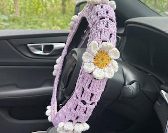 May include: A lavender crochet steering wheel cover adorned with white and yellow daisy accents. The cover is handmade with a textured open weave design, and is designed to fit a standard car steering wheel.