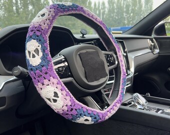 May include: A crochet steering wheel cover in shades of purple, blue, and white, featuring a skull pattern. The cover is installed on a car's steering wheel, adding a unique, handmade touch to the vehicle's interior.