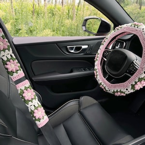 May include: Crocheted car accessories, including a steering wheel cover and seatbelt covers, featuring a floral design in pink, white, and green. The interior of a car is visible, with a black leather seat and a view of the outside.