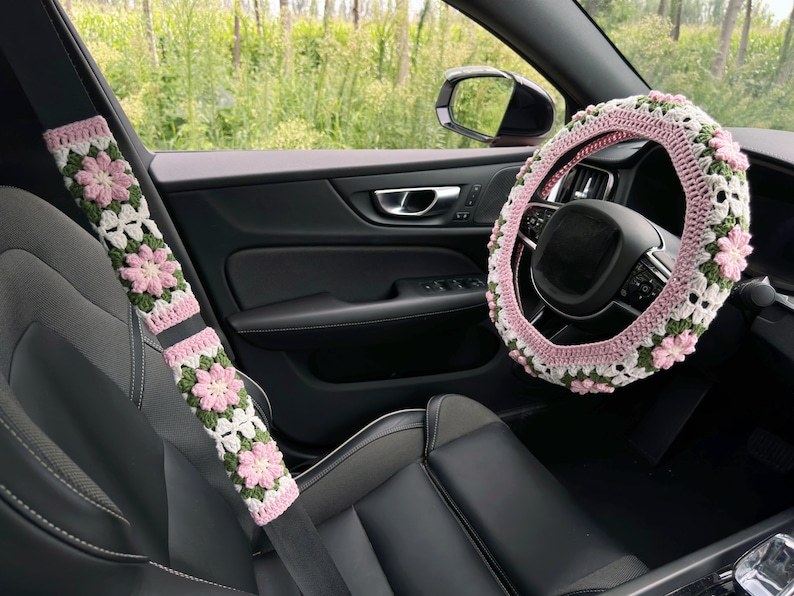 May include: Crocheted car accessories, including a steering wheel cover and seatbelt covers, featuring a floral design in pink, white, and green. The interior of a car is visible, with a black leather seat and a view of the outside.