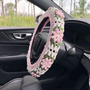 May include: A handmade crochet steering wheel cover. The cover features a pink, white, and green floral design. The cover is on a black steering wheel inside a car.