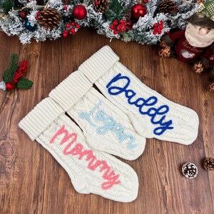 May include: Three white cable-knit Christmas stockings with embroidered names: "Mommy" in pink, "Logan" in light blue, and "Daddy" in dark blue. The stockings have a folded cuff and are displayed on a wooden surface.