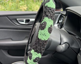 May include: A black and green crochet steering wheel cover. The cover features a repeating pattern of green and black flower-like shapes. The cover is shown on a car's steering wheel, with the interior of the car visible in the background.