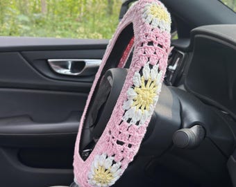 May include: A pink crochet steering wheel cover with white and yellow daisy flower designs. The cover is made of a soft, textured yarn and fits snugly around the steering wheel. The daisy flowers are evenly spaced around the cover.